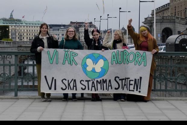 Personer från Aurora står utanför riksdagen med ett plakat. Där står det att de ska stämma staten.