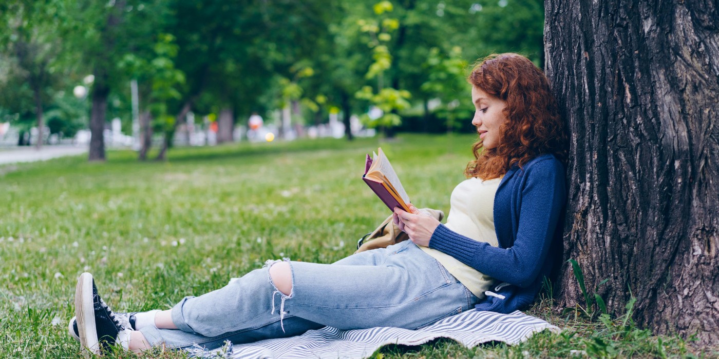 Kvinna sitter och läser en bok lutat mot ett träd.