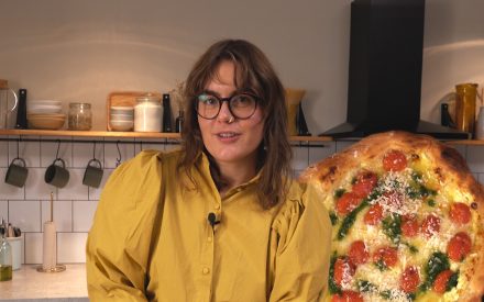 Kvinna står i gul tröja i ett kök med en pizza.