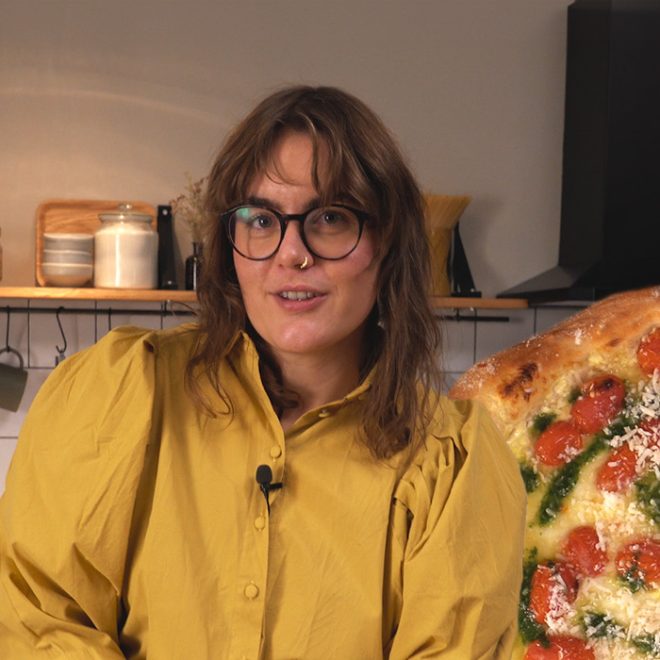 Kvinna står i gul tröja i ett kök med en pizza.