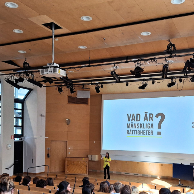 En föreläsare pratar inför en publik i en aula med en presentation om mänskliga rättigheter på skärmen.