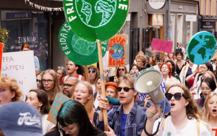 Människor går i ett demonstrationståg i centrala Stockholm. Några håller i skyltar med en grön jordglob och texten Fridays for future. Längst fram i bilden håller en kvinna i en megafon.