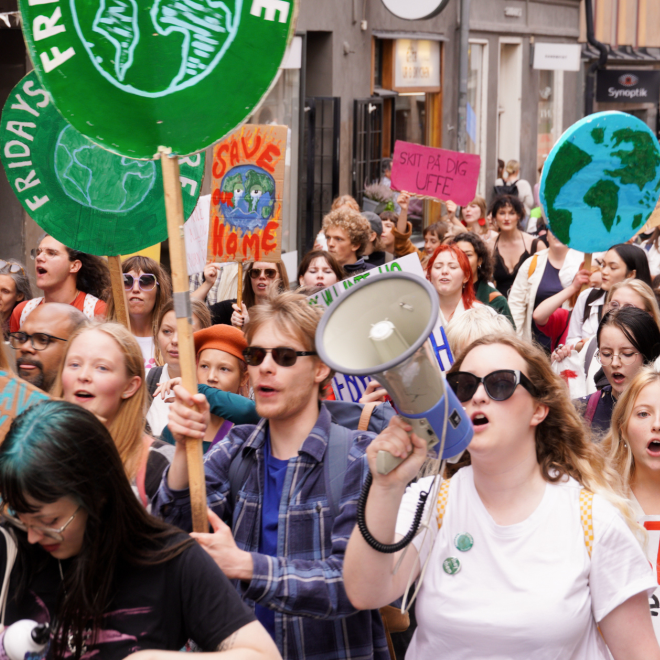 Människor går i ett demonstrationståg i centrala Stockholm. Några håller i skyltar med en grön jordglob och texten Fridays for future. Längst fram i bilden håller en kvinna i en megafon.