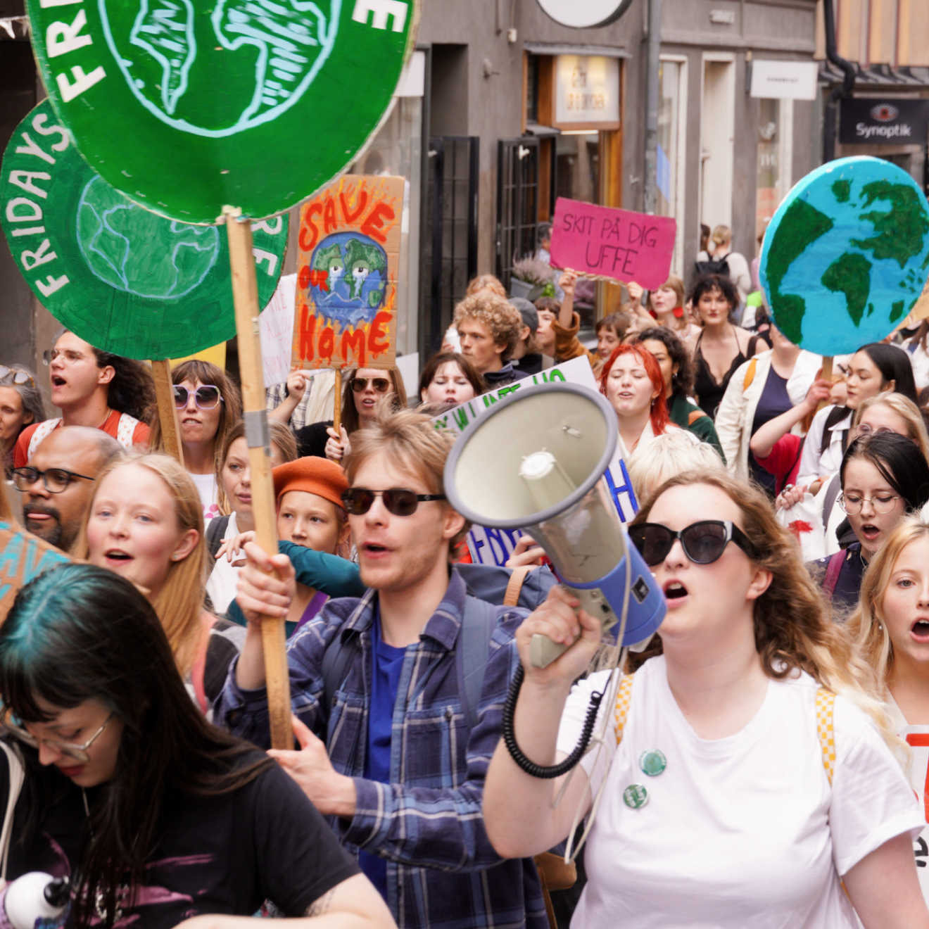 Människor går i ett demonstrationståg i centrala Stockholm. Några håller i skyltar med en grön jordglob och texten Fridays for future. Längst fram i bilden håller en kvinna i en megafon.