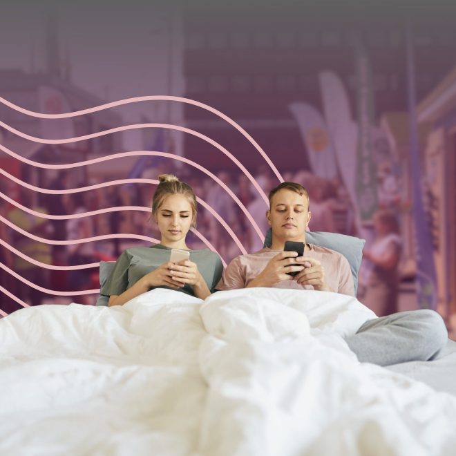 två personen sitter med sina telefoner i sängen. Bakom syns valstugor.