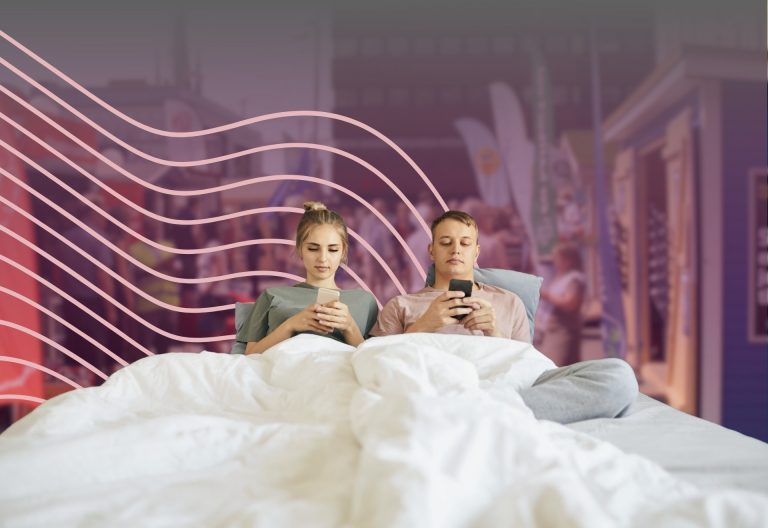 två personen sitter med sina telefoner i sängen. Bakom syns valstugor.